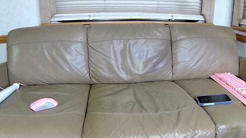 Media: Video of a beige leather couch with a pink mouse pad and a smartphone on the right armrest, set against a backdrop of white walls and a window with closed blinds.