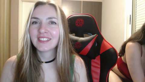 Media: Video of a young woman with long, straight blonde hair, fair skin, and a black choker, smiling. Behind her, a red and black gaming chair with a skull emblem.