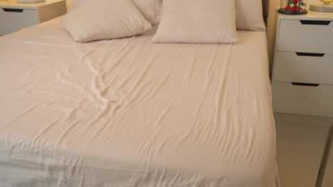 Media: Video of a neatly made bed with light beige sheets and pillows, in a minimalist, modern bedroom with white drawers and a wooden nightstand.