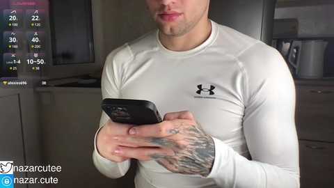 Media: Video of a white man with a fit physique and tattoos on his arms, wearing a white Under Armour shirt and a black smartphone. He stands in a modern kitchen with grey cabinets and a sink.