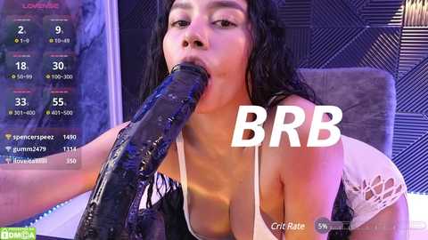 Media: A video of a woman with long black hair, wearing a white lace top, performing oral sex on a large, black dildo in a modern room.