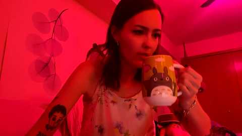 Media: A video of a woman with dark hair, wearing a floral top, drinking beer in a dimly lit room with red lighting. She has a tattoo on her left arm.