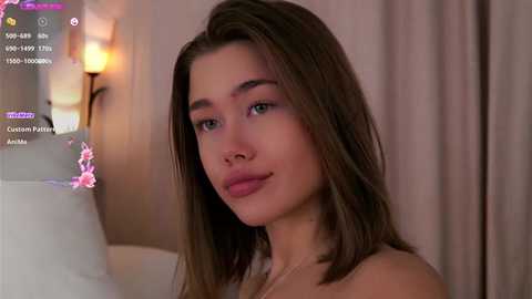 Media: Video of a young woman with light brown skin, straight brown hair, and subtle makeup, wearing a strapless top, in a softly lit, beige-toned room with a blurred background.