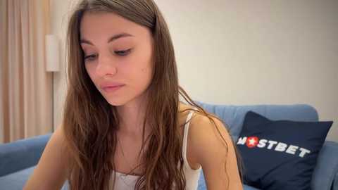 Media: Video of a young Caucasian woman with long brown hair, wearing a white tank top, sitting on a blue couch in a living room with beige walls and a \"HOSTETT\" pillow.