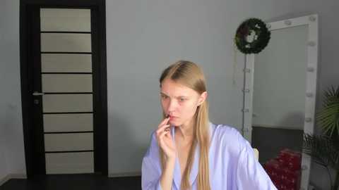 Media: Video of a young, slender, blonde woman in a white robe, holding her finger to her lips, standing in a modern, minimalist room with grey walls, black door, and a green plant.