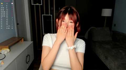 Media: Video of a young Asian woman with short red hair, covering her face with her hands, in a modern bedroom with a white desk, calendar, and bed.