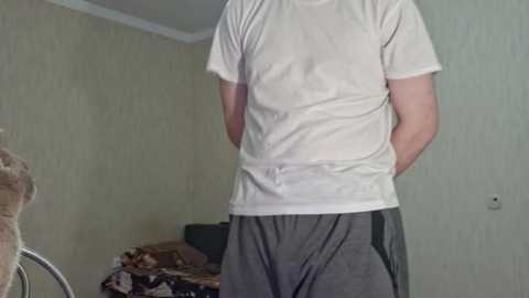 Media: Video of a person in a white T-shirt and gray sweatpants standing in a dimly lit room with beige wallpaper. A bed with a brown blanket and a small dog is visible in the background.