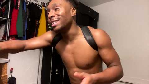 Media: Video of a muscular Black man with a wide smile, shirtless, wearing black shoulder straps, standing in a closet filled with colorful clothes.