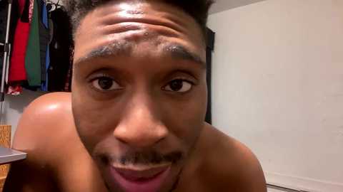 Media: Video of a shirtless Black man with a wide, mischievous grin, short curly hair, and a small goatee. Background features a hanging closet with colorful clothes.