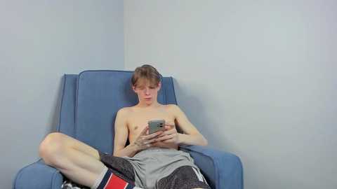 Media: A video of a shirtless, young, skinny boy with short brown hair, wearing gray shorts and red socks, sitting in a blue armchair, intensely focused on his smartphone.