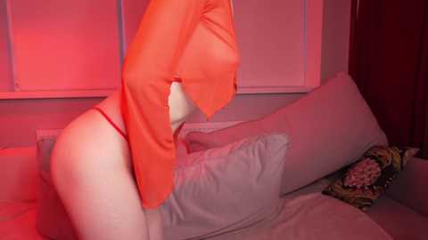 Media: Video of a woman in a red blouse, partially covering her breasts, leaning against a bed with pillows and a floral-patterned cushion, in a dimly lit room with pinkish lighting.