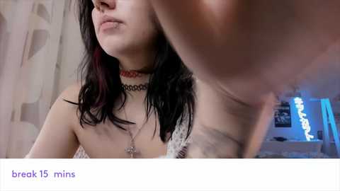 Media: Video of a pale-skinned woman with black hair, wearing a red choker and white lace bralette, looking down. Background shows a dimly lit bedroom with a blue neon sign.