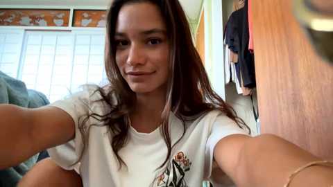 Media: Video of a smiling young woman with long brown hair, wearing a white t-shirt, taking a selfie in a room with wooden furniture, a window, and a closet.