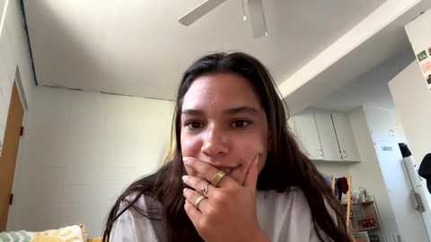 Media: Video of a young woman with long dark hair, wearing a white shirt, biting her finger, in a cluttered, brightly lit dorm room with white walls and a ceiling fan.