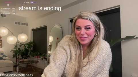 Media: Video of a smiling, blonde woman in a beige knit sweater, sitting in a modern, brightly lit living room with a large mirror, plants, and hanging lights.