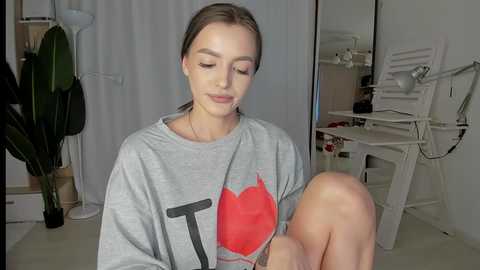 Media: Video of a young woman with fair skin, brown hair, wearing a grey sweatshirt with a red heart graphic, sitting in a minimalist room with white walls and a white chair.