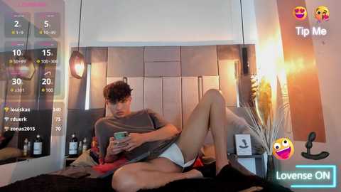 Media: Video of a young man with curly hair, wearing gray shirt and white shorts, sitting on a bed, watching a phone, in a modern bedroom with white walls, large mirror, and digital clock.