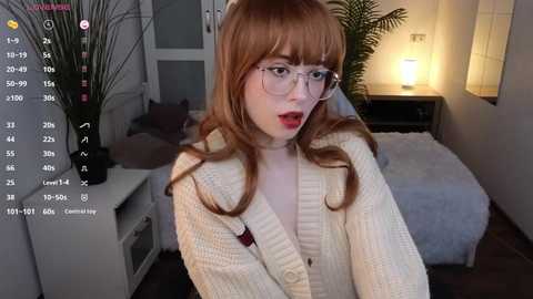 Media: A video of a pale-skinned, red-haired woman with glasses, wearing a cream sweater, in a dimly lit bedroom with a bed, potted plant, and digital clock.