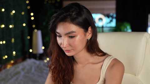 Media: Video of a young woman with long, dark hair, wearing a beige top, seated indoors near a Christmas tree with fairy lights, a beige couch, and a blurred TV screen in the background.