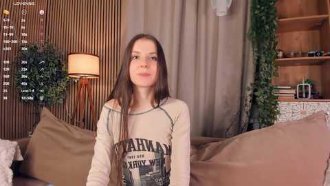 Media: Video of a young Caucasian woman with straight brown hair, wearing a beige \"Harlem World\" graphic tee, sitting on a brown couch in a cozy, wooden-paneled living room.