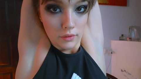 Media: Video of a young woman with fair skin and brown hair, wearing dark makeup, black halter top, in a dimly lit room with a white dresser and framed art in the background.