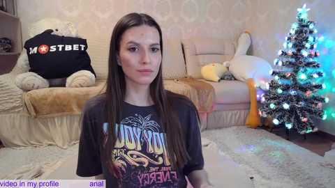 Media: Video of a young woman with long brown hair, wearing a dark t-shirt, sitting in a cozy, Christmas-decorated living room with a lit tree and plush toys.