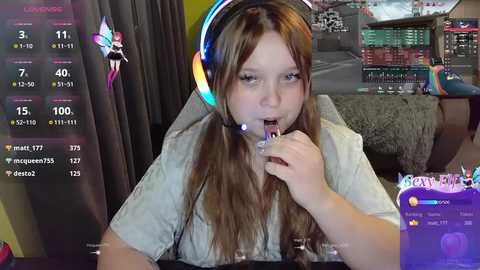 Media: Video of a young woman with long brown hair, wearing headphones, and a white shirt, playing a video game, surrounded by colorful gaming setup.