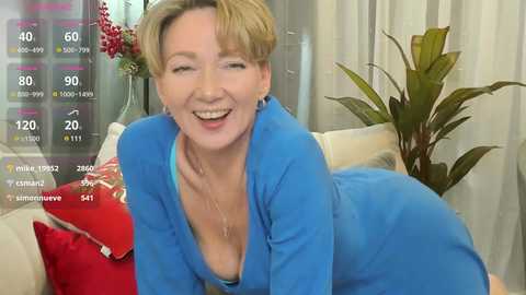 Media: Video of a smiling middle-aged woman with short blonde hair, wearing a blue dress, lying on a couch. Background includes a potted plant and red decorative pillow.