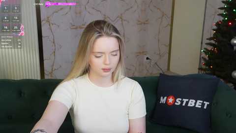 Media: Video of a fair-skinned blonde woman with straight hair, wearing a beige T-shirt, sitting on a green couch, in a room with a decorated Christmas tree and a wall with marble-like patterns.