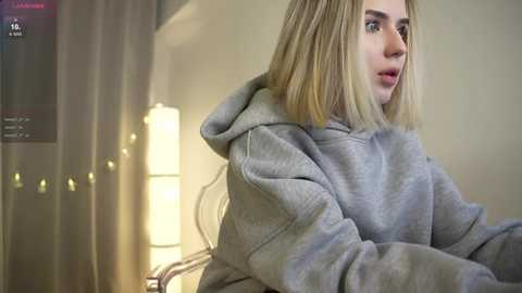 Media: A video of a young woman with shoulder-length blonde hair, wearing a grey hoodie, looking surprised in a dimly lit room with a glowing lamp and closed curtains in the background.