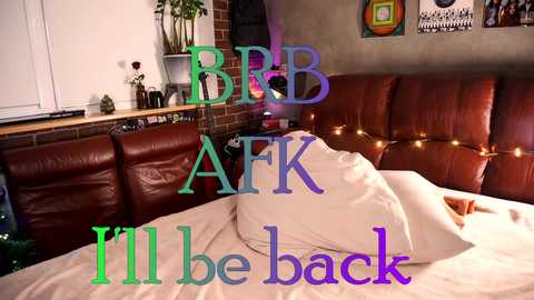 Media: Video of a cozy, dimly-lit bedroom with a brown leather couch, brick wall, and string lights; text overlay reads \"IBB AFTK I'll be back.\