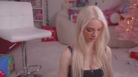 Media: Video of a young, fair-skinned woman with long, platinum blonde hair, wearing a black tank top, sitting on a beige carpet in a brightly lit, festive room with white furniture, Christmas decorations, and a pink tree.