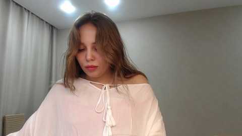 Media: Video of a young woman with straight, shoulder-length brown hair, wearing a loose white blouse with a bow tie, in a dimly lit, minimalist room with beige walls and curtains.