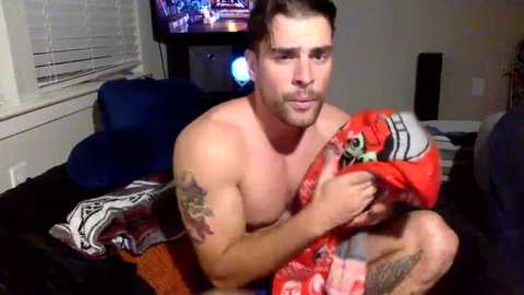 Media: Video of a shirtless, muscular, bearded man with short brown hair, sitting on a bed in a dimly lit room, holding a red and white blanket with a green toy.