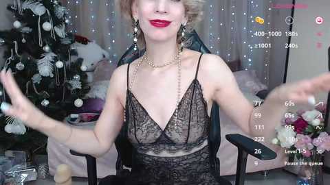 Media: Video of a blonde woman with red lipstick, wearing a black lace camisole, seated in a chair in a festive room with a decorated Christmas tree.