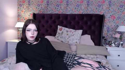 Media: Video of a young woman with pale skin and dark hair, wearing glasses and a black choker, lying on a bed with floral wallpaper, floral pillows, and a pink floral blanket.