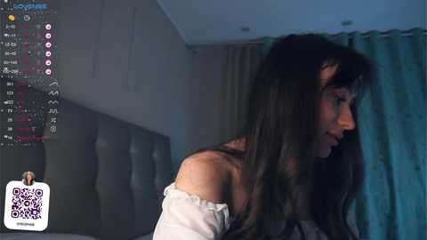 Media: A video of a young woman with long, dark hair in a dimly lit room, wearing a white off-shoulder top, captured on a Twitch stream.