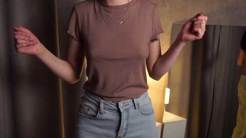 Media: Video of a woman in a fitted, short-sleeve, brown t-shirt and high-waisted, light blue jeans, standing with fists clenched in a dimly lit, beige-walled room with a mirror and lamp in the background.