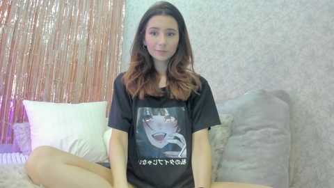 Media: A video of a young woman with long brown hair, wearing a black graphic t-shirt featuring a cartoon dog, sitting on a bed with white and beige pillows, against a textured wall and metallic curtain backdrop.