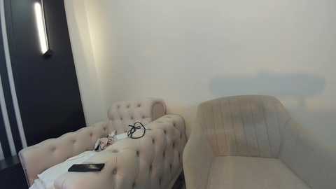 Media: Video of a small, sparsely furnished room with beige tufted sofas, a black wall, and a modern light fixture. A smartphone and headphones are on one sofa. The room appears dimly lit.