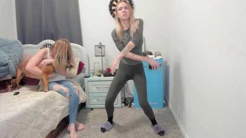 Media: Video of a blonde woman in a green onesie, dancing energetically in a cluttered, beige-carpeted bedroom with a bed, dresser, and a blue bin.