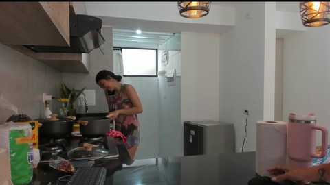 Media: Video of a woman in a modern, minimalist kitchen with white walls and stainless steel appliances. She's cooking, wearing a colorful apron, and the kitchen is cluttered with items on the counter.