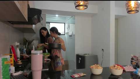 Media: Video of two women in modern kitchen preparing food; one in a tank top, the other in a sleeveless top, using a blender; countertop cluttered with bowls, utensils, and condiments.