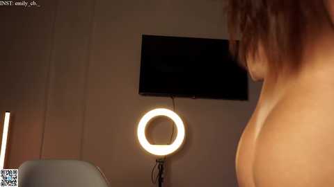 Media: Video of a nude woman with light skin, shoulder-length brown hair, standing in a dimly lit room with a circular light and a flat-screen TV on the wall.