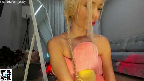 Media: Video of a young, fair-skinned woman with long, blonde hair styled in a braid, wearing a pink strapless top, standing in a modern, dimly lit living room with a gray couch and a mirror reflecting the room.