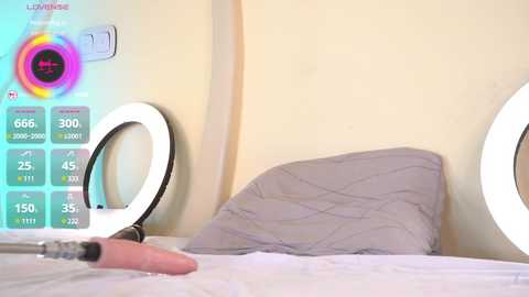 Media: Video of a minimalist bedroom with a gray bed, a pink vibrator on the pillow, and a colorful light ring on the wall, displaying a thermometer and a calendar.