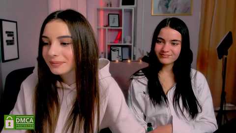 Media: Video of two young women with long dark hair, wearing white shirts, seated in a dimly lit room with bookshelves, framed pictures, and a spotlight.