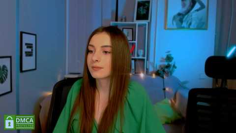 Media: Video of a young woman with long brown hair in a green top, seated indoors. Background shows a modern living room with bookshelves, framed art, and a dimly lit scene.