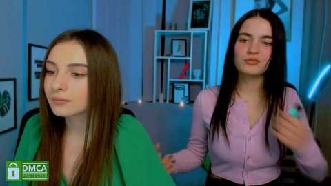 Media: Video of two young women in a modern, dimly lit room. One has long brown hair, wearing a green shirt, looking sad. The other, with long black hair, wears a lavender cardigan and a smile.