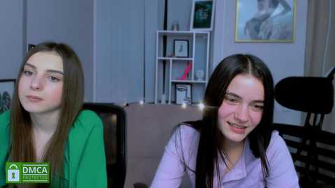 Media: Video of two young women with light skin, long dark hair, and fair complexions. They are seated, smiling, wearing green and lavender tops. Background shows a bookshelf, framed artwork, and a dark chair.